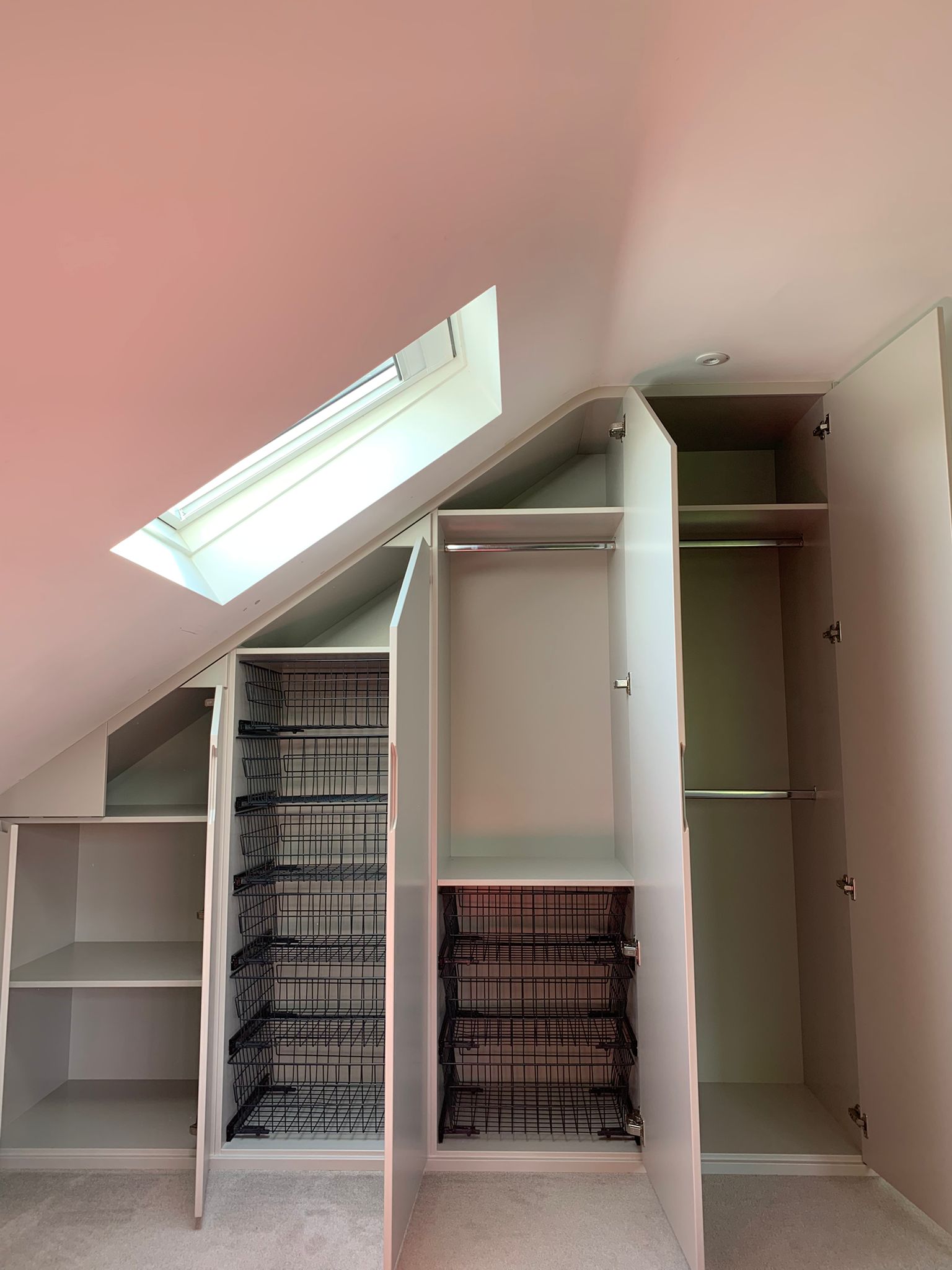 23667 Interior of a bespoke loft wardrobe with sloped ceiling design, featuring hanging rails, pull-out storage baskets, shelving, and custom-fitted compartments built into an attic bedroom in Dorset.