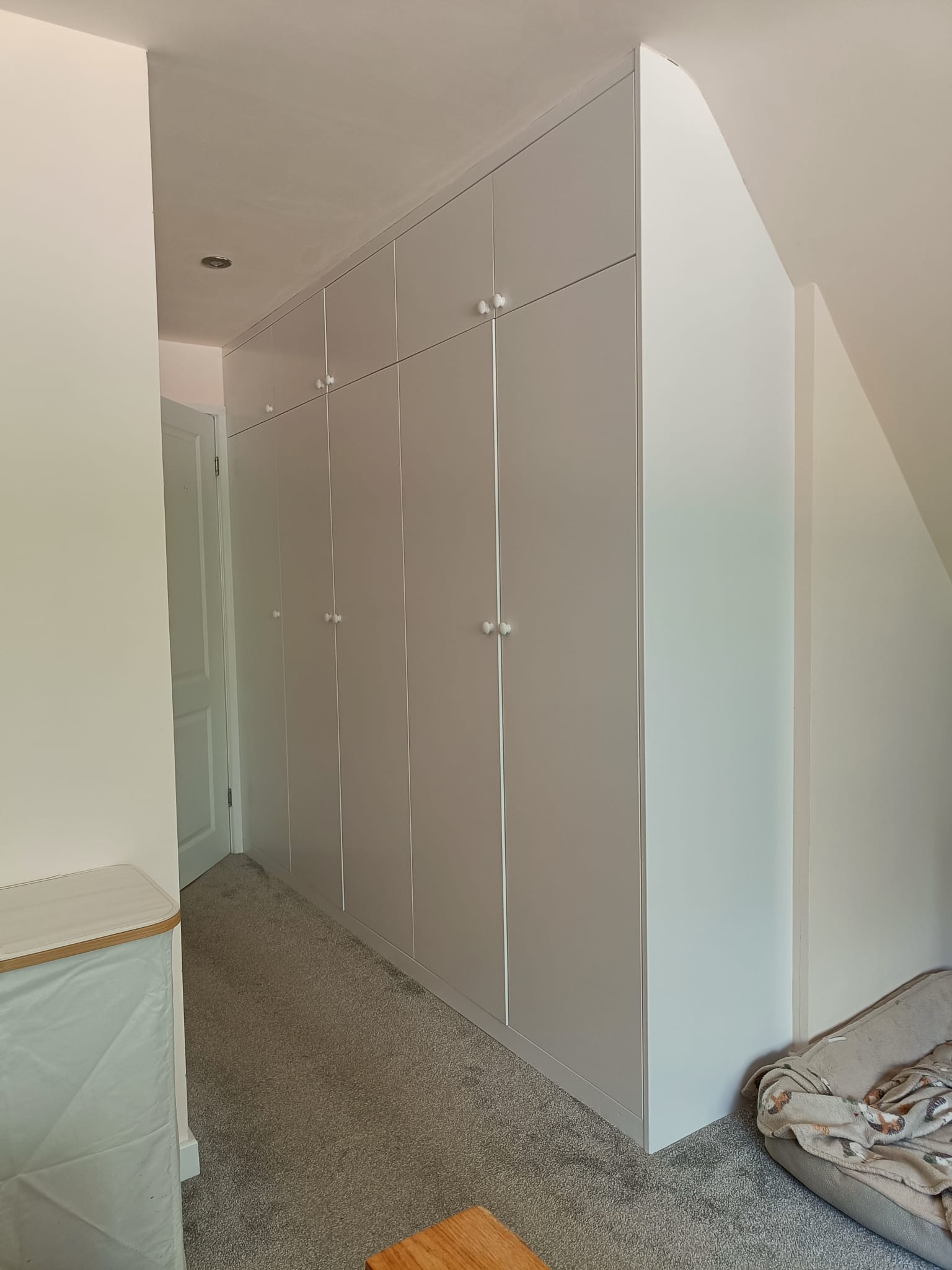 IMG-20260502-WA0018(1) Bespoke fitted wardrobe built into a sloped loft space with floor-to-ceiling shaker-style doors, overhead storage cupboards, and custom scribed panels for a seamless fitted finish in a Dorset home.
